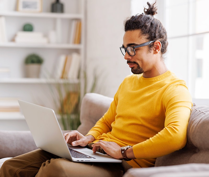 Un homme portant des lunettes et une chemise jaune, assis sur un canapé avec un ordinateur portable, plongé dans son travail et se demandant si son Mac peut être infecté par des virus.