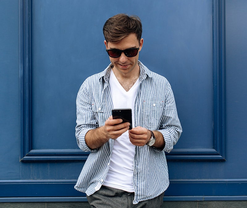 Un jeune homme portant des lunettes de soleil consulte son smartphone, cherchant comment supprimer son compte Facebook.