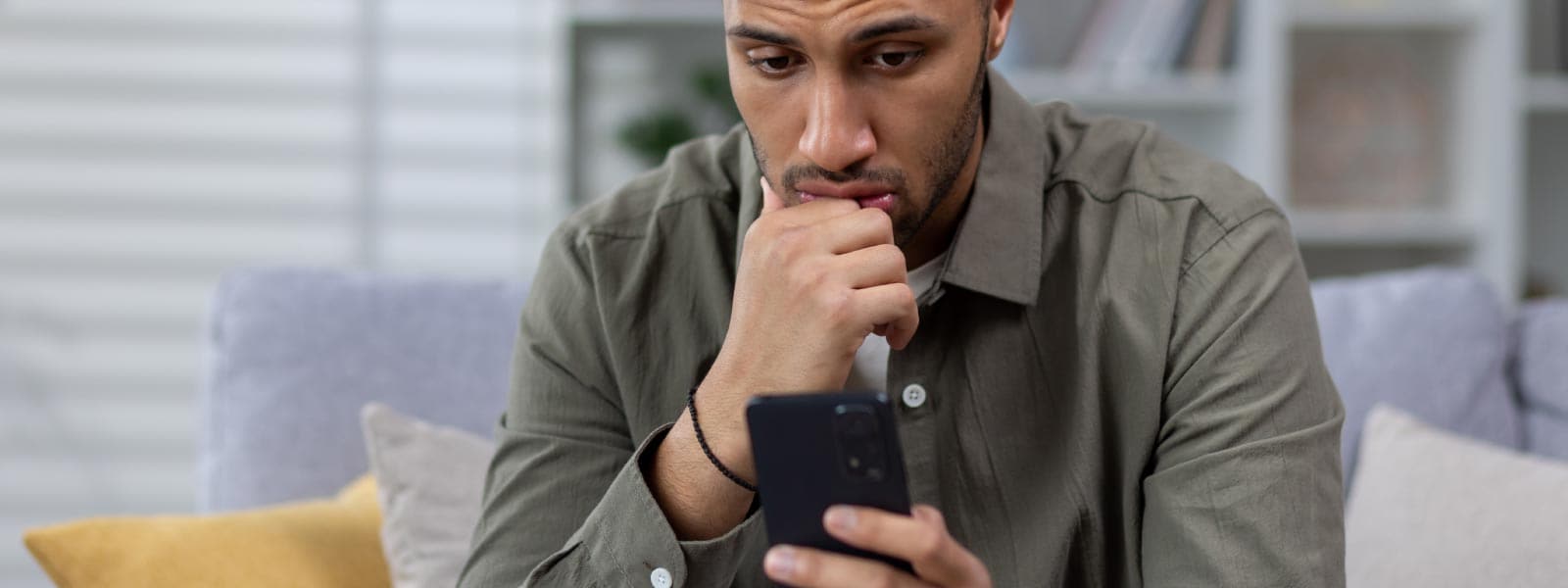 Un homme inquiet, vêtu d’une chemise grise, regarde son téléphone, ne sachant pas s’il est sur écoute ou a été piraté.