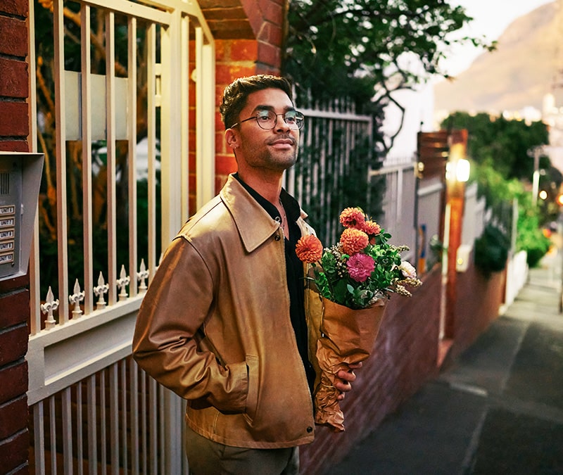 Un homme attend devant une maison en briques, un bouquet de fleurs à la main.