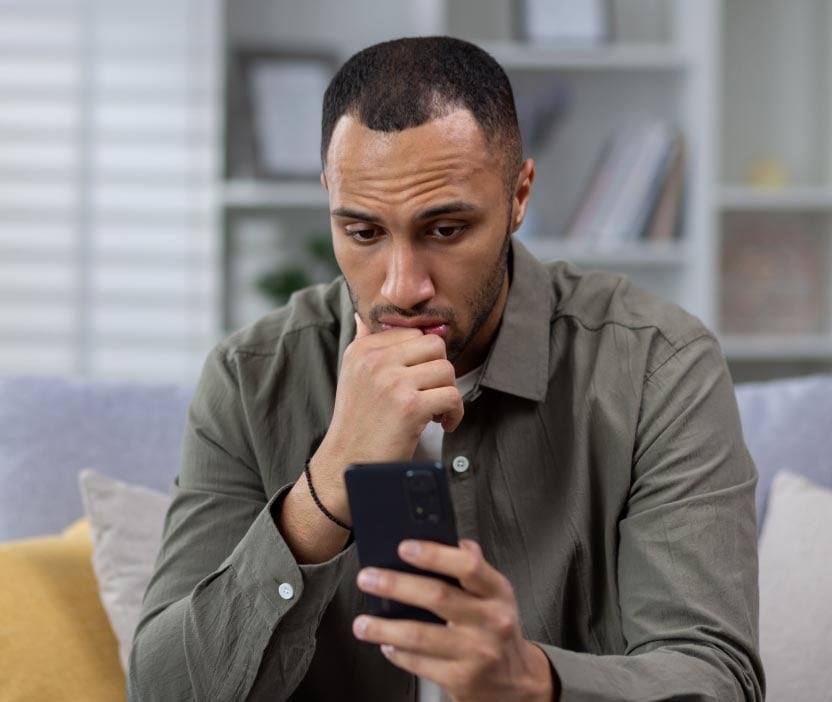 Un homme inquiet, vêtu d’une chemise grise, regarde son téléphone, ne sachant pas s’il est sur écoute ou a été piraté.