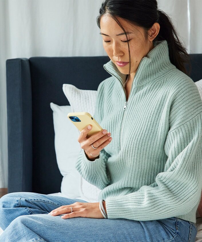 Une femme assise sur un lit, regarde son téléphone et réfléchit à la façon de créer un mot de passe mémorable.