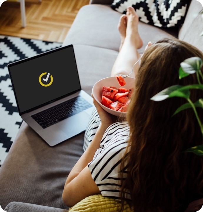 Femme mangeant des fruits avec un ordinateur portable affichant le logo NortonLifeLock posé à côté d'elle.