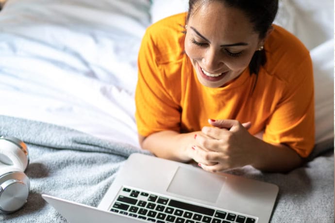 Une femme souriante couchée sur un lit qui utilise un ordinateur portable.